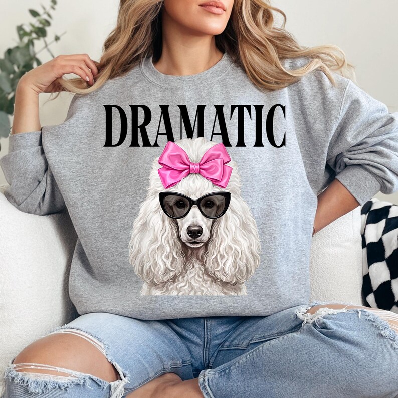 May include: A gray sweatshirt with the word "DRAMATIC" in black block letters. The sweatshirt features a graphic of a white poodle wearing black sunglasses and a pink bow. The poodle's fur is detailed and fluffy.