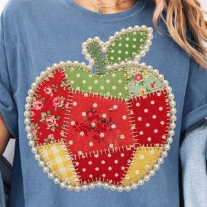 May include: A light blue t-shirt featuring a patchwork apple design. The apple is composed of various fabric patterns, including red floral, polka dots, and yellow gingham. The apple is outlined with a pearl-like trim, creating a decorative effect.