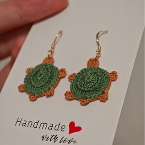 May include: Handmade crochet turtle earrings. The earrings have green turtle shells with orange edging and gold-coloured ear wires. The earrings are displayed on a white card with the text "Handmade with love" and a red heart.