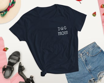 May include: Navy blue t-shirt with the embroidered text "Dog Mom" and a paw print design. The shirt is styled with denim shorts, black sandals, a straw hat, and red sunglasses. Strawberries are scattered around the items.