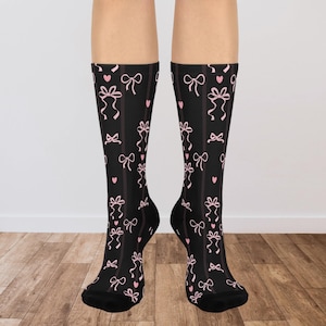May include: Black knee-high socks with a pattern of pink bows and hearts. The socks have vertical stripes and a repeating design of bows and hearts. The socks are on a person's legs.
