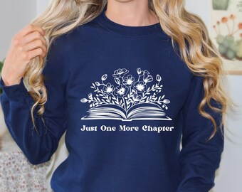 Peut inclure: Un sweat-shirt bleu marine avec un motif blanc d'un livre ouvert avec des fleurs sur le dessus. Le texte "Just One More Chapter" est imprimé sous le livre.