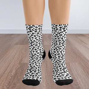 May include: A pair of crew socks with a white background and a black leopard print pattern. The socks have black heels and toes. The socks are displayed on a wooden floor.