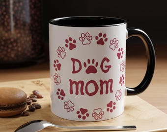 Könnte beinhalten: Ein weißer Keramikbecher mit schwarzem Innenraum und Henkel. Die Außenseite zeigt den Schriftzug "Dog mom" in roten Buchstaben, umgeben von einem kreisförmigen Muster aus roten Pfotenabdrücken. Das Design ist mittig platziert.