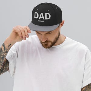Könnte beinhalten: Eine schwarze Snapback-Cap mit grauem Schirm und einem grauen Knopf an der Oberseite. Auf der Vorderseite der Kappe befindet sich der weiße Schriftzug DAD in einer serifenlosen Schrift, mit dem kleineren weißen Text est 2026 darunter.