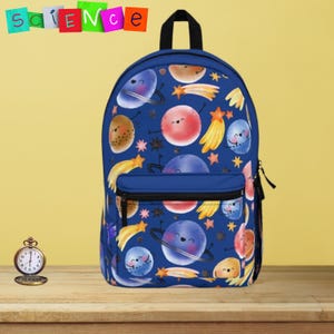 May include: A navy blue backpack featuring a repeating pattern of smiling planets, stars, and comets. The backpack has a black handle and a front pocket. The word "science" is spelled out in colorful letters at the top.