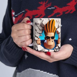 Puede incluir: Taza de cerámica blanca con un diseño de personaje de dibujos animados. El personaje, con pelo naranja y gafas de sol, parece estar rompiendo una pared gris. La taza la sostiene una persona que lleva un jersey azul marino.