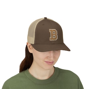Op de afbeelding: Bruine truckerpet met een beige mesh achterkant en een bruine klep. De pet heeft een groot, geborduurd "B" in een lichtere bruine kleur op de voorkant. Gedragen door een persoon met een groen shirt.