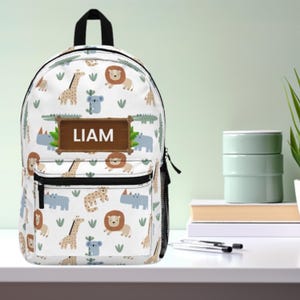 May include: White backpack with a black handle and zipper, featuring a repeating pattern of cartoon animals including lions, giraffes, and rhinos. A brown rectangular patch displays the name "LIAM". The backpack has a front pocket and a side mesh pocket.