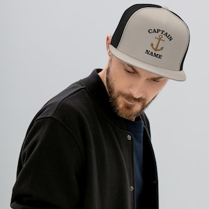 May include: A black and beige trucker hat with a gold anchor and the text "CAPTAIN NAME" embroidered on the front.