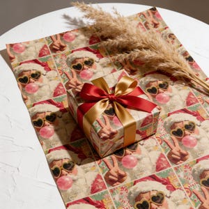 May include: A square gift box wrapped in paper featuring a repeating pattern of Santa Claus wearing heart-shaped sunglasses and blowing a pink bubble, tied with a shiny red and gold ribbon. A dried pampas grass stem rests on the wrapping paper.
