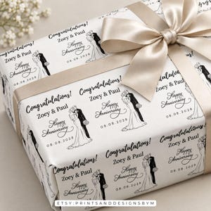 Op de afbeelding: Een witte geschenkdoos verpakt in papier met een herhalende zwarte lijntekening van een koppel, de tekst Congratulations! Zoey & Paul, Happy Anniversary en de datum 08.08.2028, afgewerkt met een brede, gladde champagnekleurige satijnen strik.