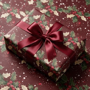 May include: A rectangular gift box wrapped in dark burgundy paper featuring a pattern of green holly leaves, red berries, and white snow-like speckles. The box is tied with a smooth, dark burgundy satin ribbon tied into a centred bow.