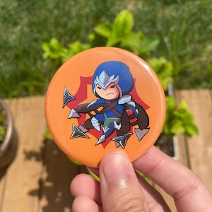 May include: A round orange button with a cartoon character in blue and brown clothing, with stylized arrowheads around it. The character has a stern expression. The button is held in a hand against a blurred green background.