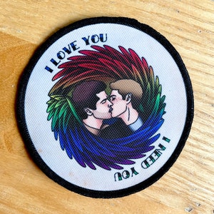 May include: Round patch with a black border featuring two men kissing, surrounded by rainbow-colored wings. The words "I LOVE YOU" and "I NEED YOU" are printed in teal around the image. The patch is on a light-colored surface.