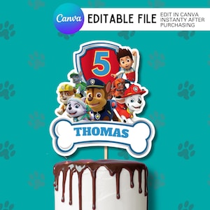 May include: A cake topper featuring the Paw Patrol characters, a red shield with the number 5, and a bone-shaped banner that says "Thomas". The cake is white with chocolate icing dripping down the sides. The background is teal.