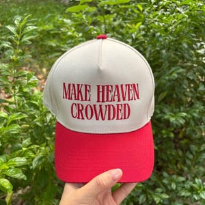 Peut inclure: Une casquette de baseball bicolore avec une calotte beige clair et une visière rouge unie. L'avant de la calotte présente le texte rouge brodé "MAKE HEAVEN CROWDED" dans une police serif grasse. La casquette a une structure à cinq panneaux.