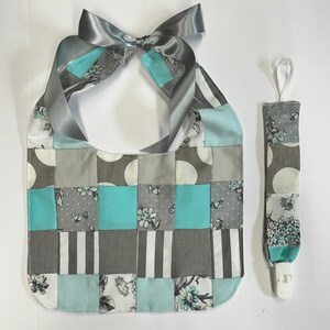 May include: A patchwork baby bib and pacifier clip set. The bib features a square pattern in shades of gray, teal, and white, with floral and polka dot accents. A gray satin ribbon forms the neck tie. The pacifier clip mirrors the bib's design.