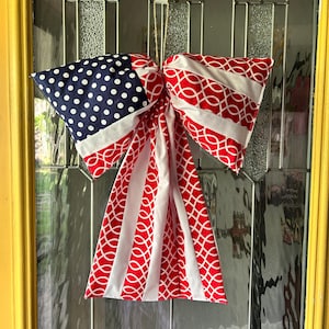 May include: A patriotic door bow featuring a design reminiscent of the American flag. The bow has a blue section with white polka dots, red and white striped sections, and red and white patterned sections. It is hanging on a glass door.
