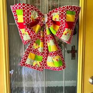 May include: A large, decorative bow made of red fabric with a white geometric pattern. The bow features patches of yellow, green, and cherry-printed fabric. It is hanging on a glass door, adding a festive touch to the entrance.