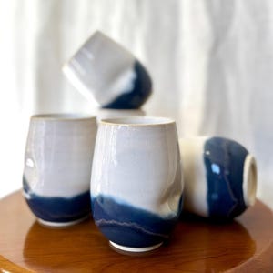 May include: Four stemless ceramic wine glasses. The glasses are white with a dark blue wave design at the bottom. One glass is lying on its side. The glasses are on a wooden surface.