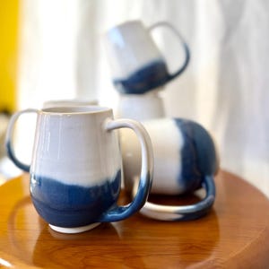 May include: A collection of ceramic mugs with a two-tone design. The mugs are white at the top, transitioning to a deep blue at the base. Each mug has a large, curved handle. The mugs are arranged on a wooden surface.