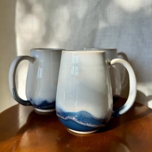 May include: Three ceramic mugs with a white and blue gradient design. The mugs have a rounded shape with a large handle and a dark blue band at the base. The mugs are sitting on a wooden surface.