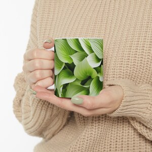 Peut inclure: Mug en céramique blanche avec un motif de feuilles de hosta vertes et blanches. Les feuilles présentent une texture et des nervures visibles. La tasse est tenue par une personne portant un pull beige.