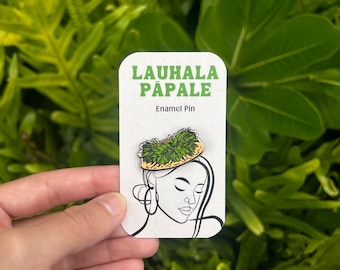 May include: A rectangular enamel pin with the text "LAUHALA PĀPALE" in green and the words "Enamel Pin" below. The pin features a line drawing of a woman wearing a hat made of leaves. The background is lush green foliage.