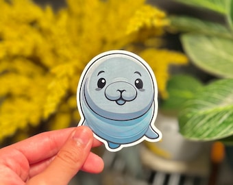 May include: A cartoon seal sticker with a light blue and white body and a light blue face. The seal has big, round eyes, a small nose, and a big smile. The sticker is perfect for adding a touch of whimsy to any project.