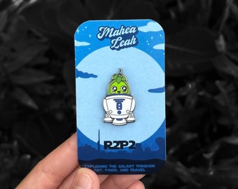 May include: A blue and white enamel pin featuring a green character with a smiling face inside a white robot. The pin is on a card with the text "Mahea Leah" and "R2P2" and the phrase "Exploring the galaxy through art, food, and travel."