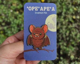May include: A brown enamel pin featuring a cartoon bat with red ears and wings, perched on a branch. The pin is on a blue card with the text "'OPE'APE'A Enamel Pin" and a full moon graphic.