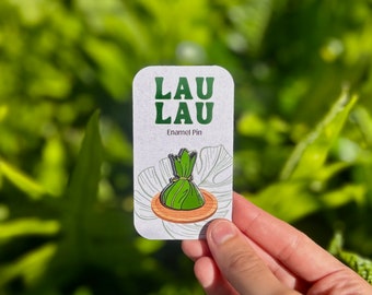 May include: A green Lau Lau enamel pin on a white card with the text "Lau Lau Enamel Pin".