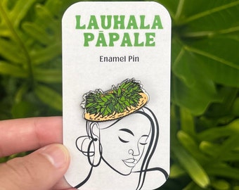 May include: A green and yellow enamel pin featuring a Lauhala Papale design. The pin is on a white card with the words "LAUHALA PĀPALE" in green and "Enamel Pin" below. A line drawing of a woman's face is on the card.