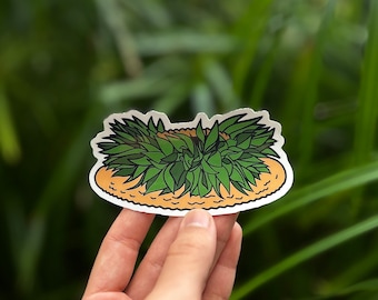 May include: A green and brown sticker of a pineapple with a straw hat on top. The sticker is being held in a hand against a green background.