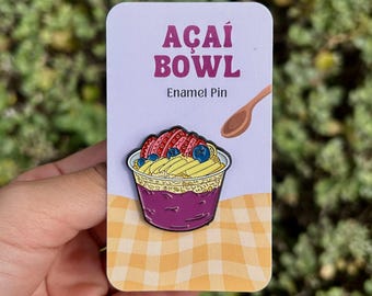 May include: Enamel pin featuring an açai bowl with strawberries, blueberries, and bananas. The pin is on a card with the words "AÇAI BOWL" and "Enamel Pin". The background is a yellow and white checkered pattern.