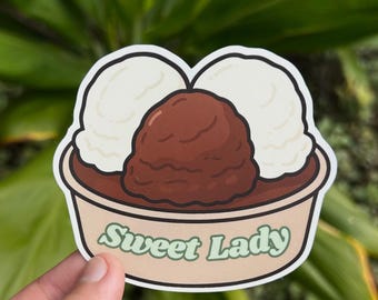 May include: A sticker featuring a dessert with two scoops of white ice cream and a scoop of chocolate ice cream in a bowl. The text "Sweet Lady" is written in green cursive font on the bowl.
