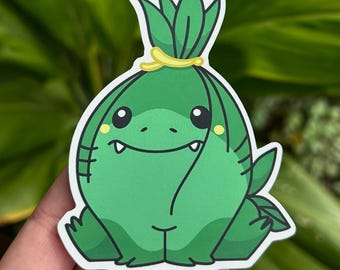 May include: A green sticker featuring a cartoon character with a round body, small fangs, and a smiling expression. The character has a leafy top and is secured with a yellow band. The sticker is held against a blurred green background.