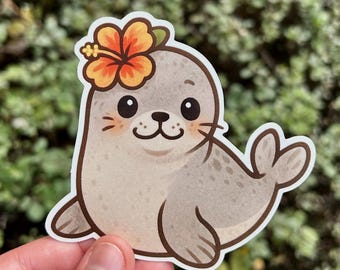 May include: A sticker featuring a cartoon seal with a flower in its hair. The seal is light brown with darker spots and a smiling face. The flower is orange and yellow. The sticker is held in a hand against a green background.