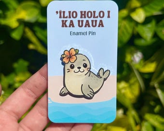 May include: A light blue enamel pin featuring a cartoon seal with a flower in its hair, set against a beach and ocean backdrop. The pin has text that reads "'ILIO HOLO I KA UAUA" and "Enamel Pin".