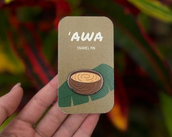 May include: A brown enamel pin with a white outline depicting a wooden bowl filled with a brown liquid. The pin is on a brown card with green leaves and the text 'AWA ENAMEL PIN'.