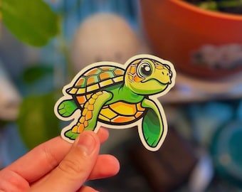 May include: A colorful sticker featuring a cartoon sea turtle. The turtle has a green head and flippers, a yellow and orange shell, and large, expressive eyes. The sticker is held by a hand.