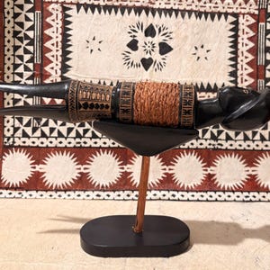 Puede incluir: Un artefacto de madera negra tallada con una sección central marrón tejida, exhibido sobre un soporte negro con un soporte de madera. El artefacto tiene forma de pájaro con extremos puntiagudos y detalles tallados intrincados. El fondo presenta un textil estampado.