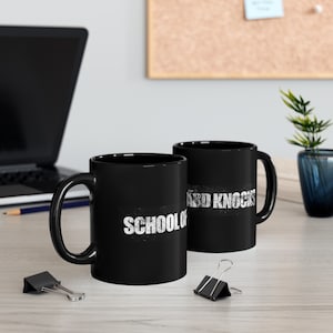 Könnte beinhalten: Zwei schwarze Kaffeetassen mit weißem Text. Die Tasse links sagt "School of Hard Knocks". Die Tasse rechts sagt "Hard Knocks".