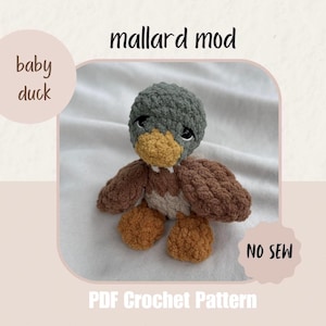 May include: A crochet mallard baby duck toy with a dark gray head, yellow beak, brown wings, and orange feet. The image includes the text mallard mod, baby duck, NO SEW, and PDF Crochet Pattern. The toy is made of soft, textured yarn.