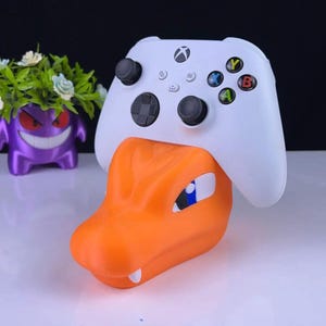May include: An orange and white controller stand shaped like a cartoon dragon's head, holding a white Xbox controller. The stand has blue and white accents. A purple cartoon character is in the background.