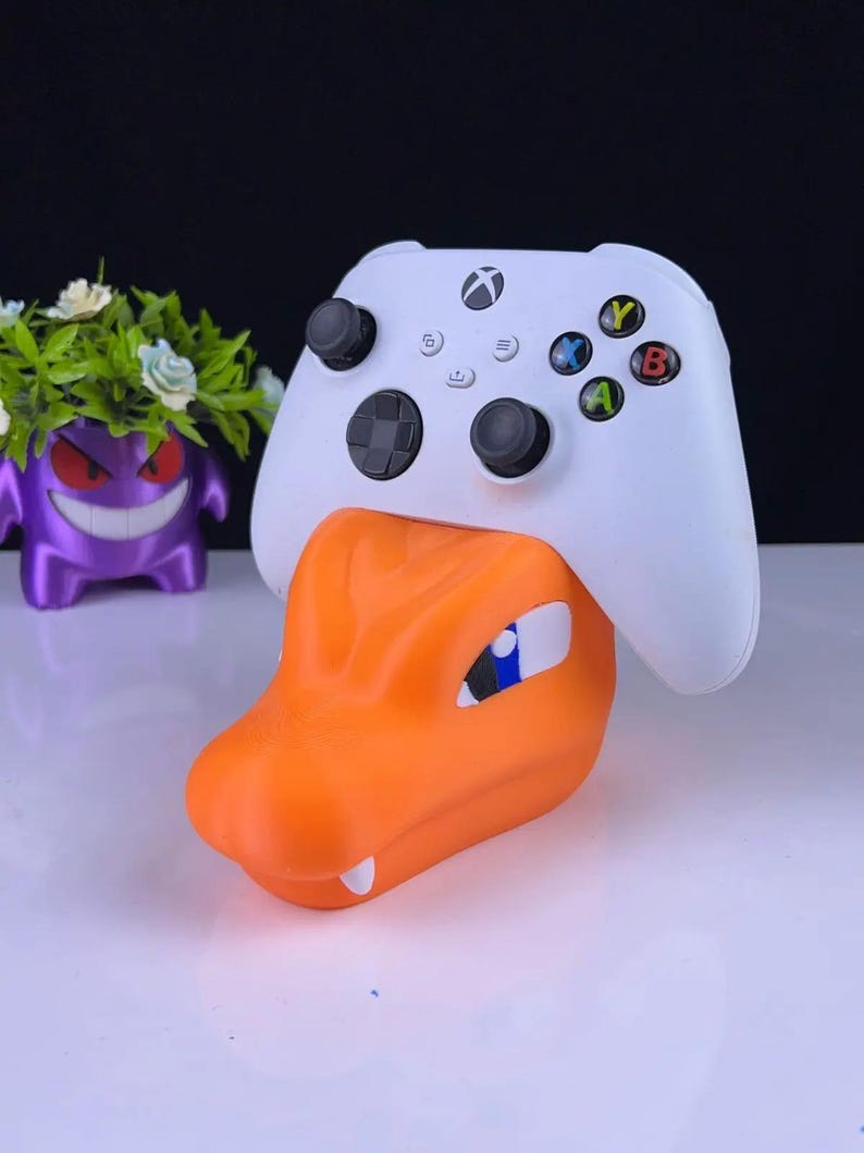 May include: An orange and white controller stand shaped like a cartoon dragon's head, holding a white Xbox controller. The stand has blue and white accents. A purple cartoon character is in the background.