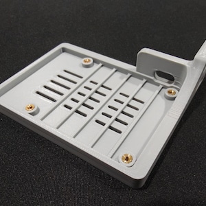 May include: A light gray rectangular plastic component with ventilation slots and brass-colored threaded inserts. The component has a raised edge and a right-angled extension with a hole.