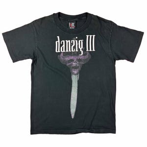 May include: Black t-shirt featuring the text "danzig III" above a graphic of a skull with a long, sword-like design. The shirt has a crew neck and short sleeves. The brand tag is visible at the top.