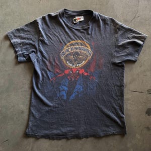 May include: Dark gray t-shirt featuring a circular graphic with the text "BLACK SABBATH WORLD TOUR". The graphic includes a skull and a red and blue devil-like figure. The shirt has short sleeves and a crew neck.